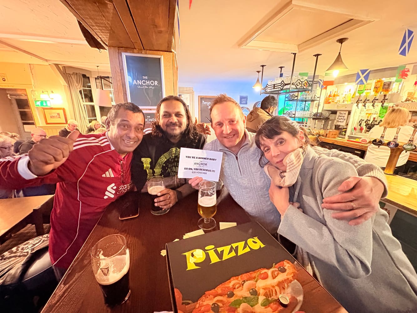 Friends holding a The Anchor voucher with pizza and drinks on the table